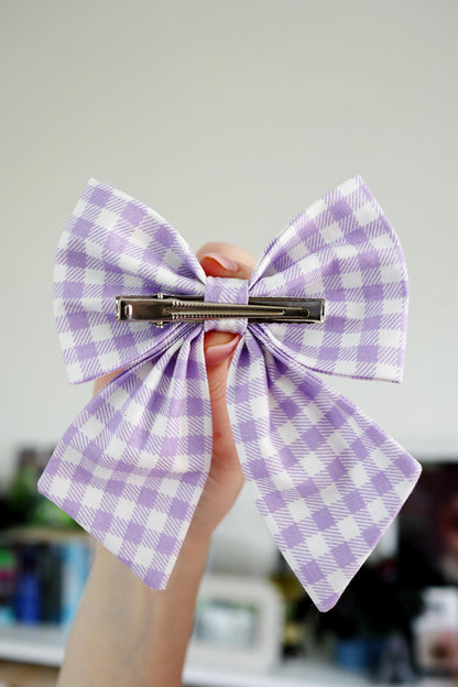 Purple and white checkered bow hair clip held by a hand against a blurred indoor background