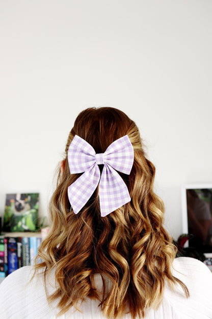 Person with wavy hair wearing a small purple and white checkered bow in the back against a white wall.