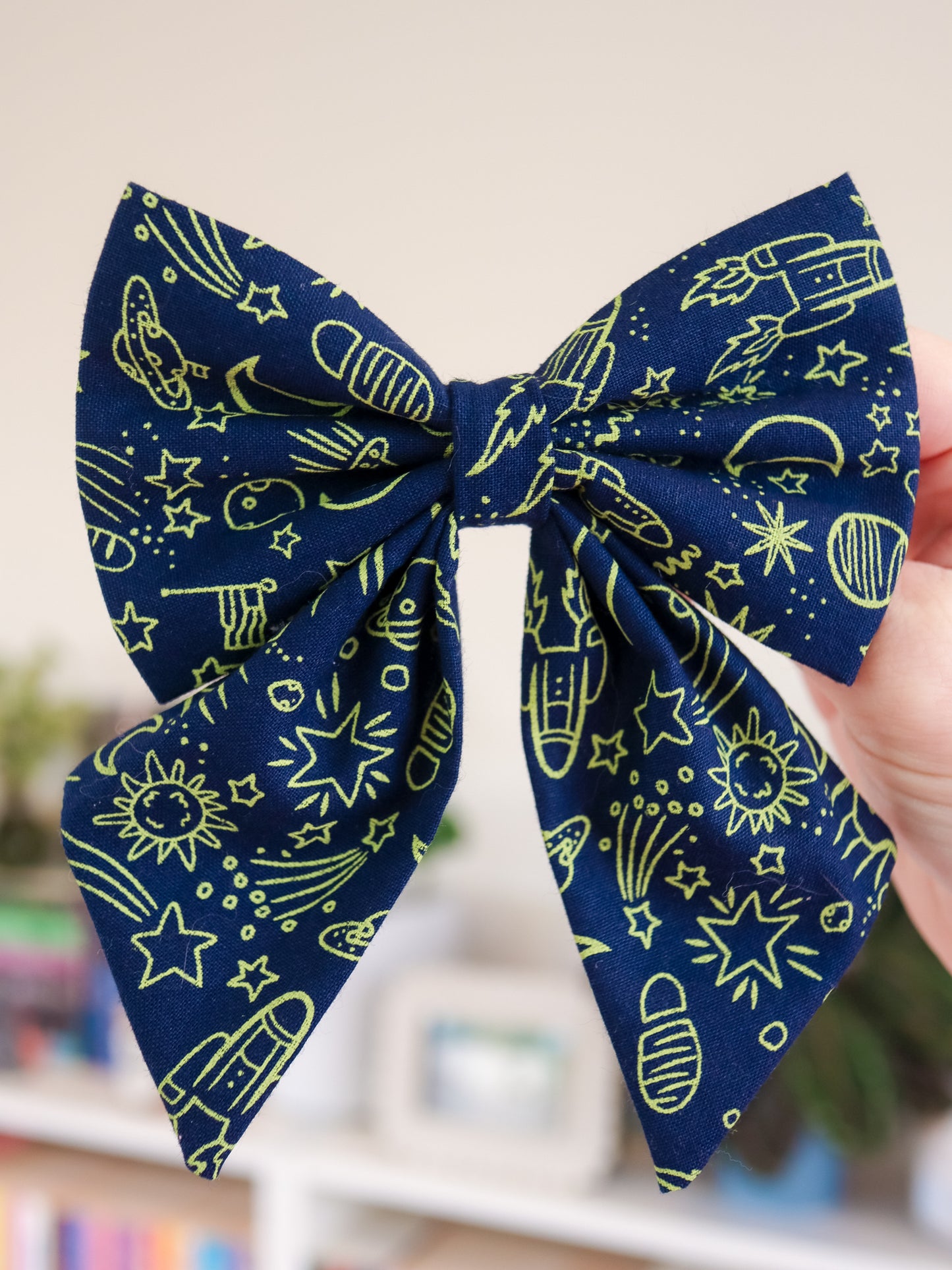 Navy blue bow with neon green space-themed pattern held by a hand against a blurred indoor background.