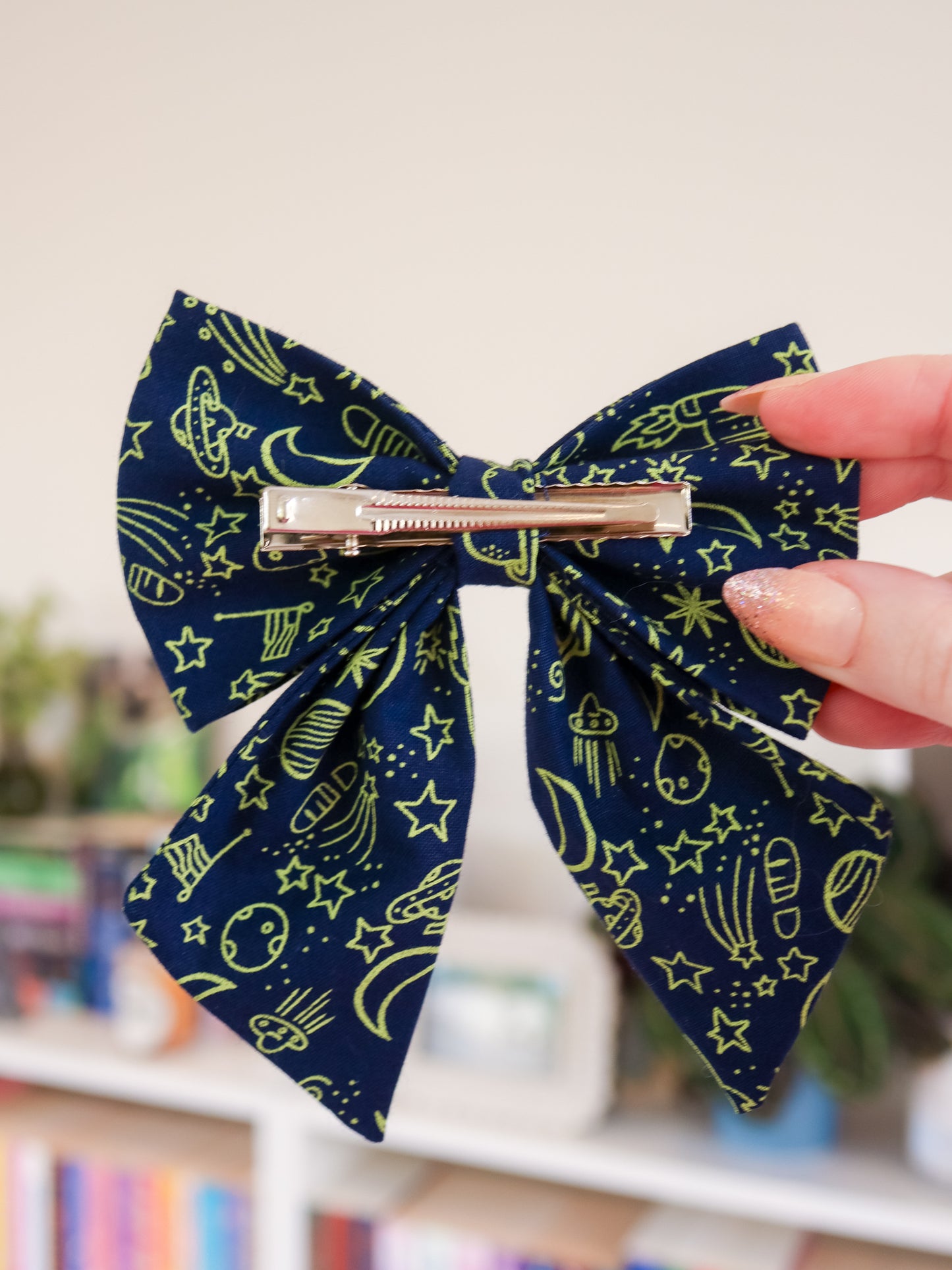 Navy blue bow with neon green space-themed pattern held by a hand against a blurred indoor background, showing alligator clip attachment