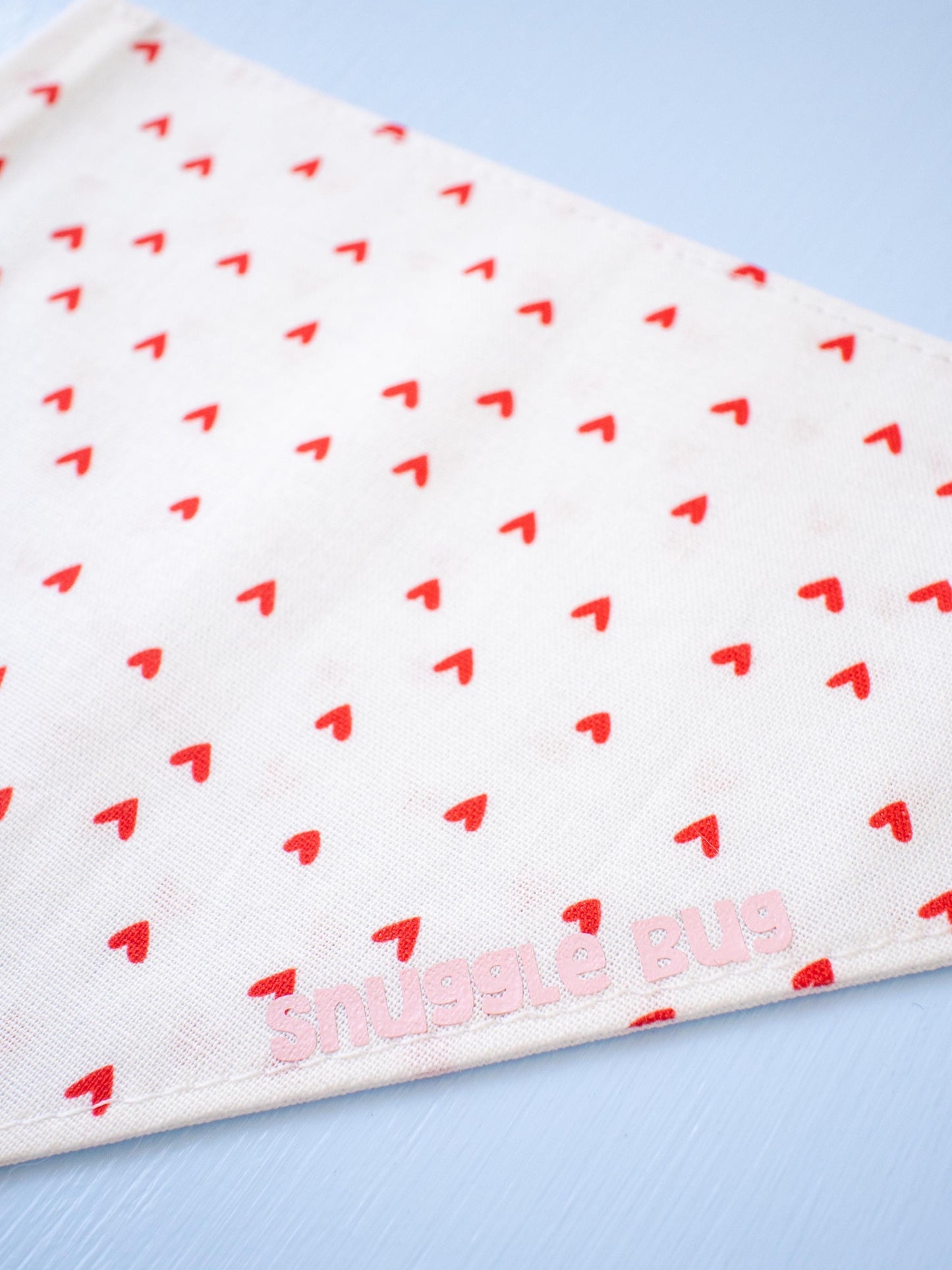 White dog bandana with red heart pattern and 'Snuggle Bug' branding on a light blue background
