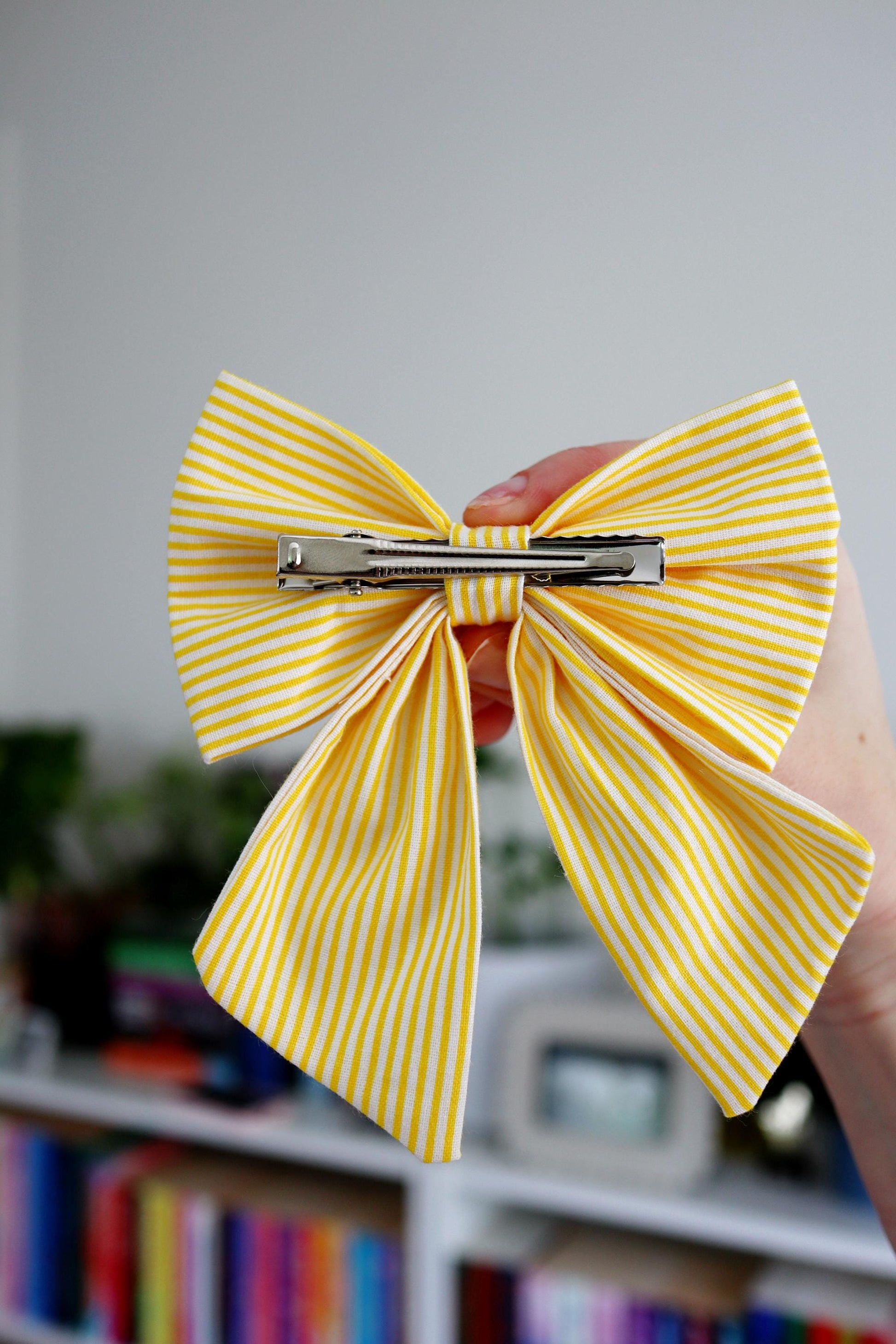 Yellow striped hair bow held by a hand with a blurred bookshelf background