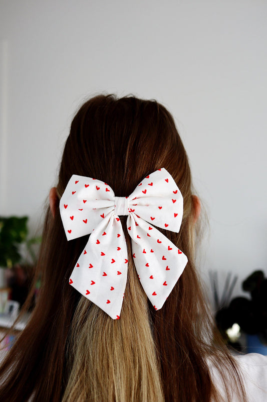 Person with a large white hair bow with red heart patterns on a blurred background